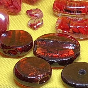 May include: A collection of red and brown glass beads. The beads are various shapes and sizes, including round, oval, and rectangular. Some of the beads have a shiny, iridescent finish.
