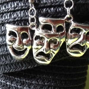 Op de afbeelding: Zilverkleurige oorbellen met drie kleine, gedetailleerde theatermaskers. De maskers zijn een lachend gezicht, een huilend gezicht en een neutraal gezicht.
