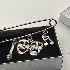 Puede incluir: Un imperdible plateado con colgantes: un micrófono, máscaras de comedia y tragedia, y una nota musical. El imperdible descansa sobre una superficie negra dentro de una caja de regalo plateada, con temática de artes escénicas.