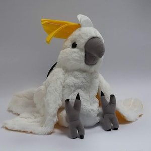 May include: White plush cockatoo stuffed animal with a yellow crest and grey feet. The cockatoo is sitting with its wings spread out.