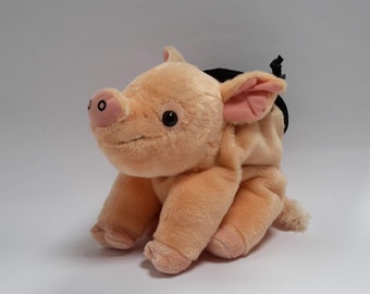 Pode incluir: Um porco de pelúcia em posição sentada. O porco é pêssego claro com focinho, orelhas e pés rosa. Tem olhos pretos e uma pequena cauda encaracolada. O porco tem uma bolsa preta nas costas.