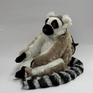 May include: A plush toy lemur with a long, striped tail. The lemur is sitting with its legs crossed and has a white and grey fur pattern. It has black eyes with orange pupils.