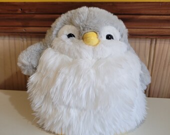 Pode incluir: Um brinquedo de pelúcia de pinguim com um corpo branco e cinza e um bico amarelo. O pinguim está sentado em uma superfície branca.