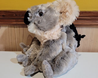 Pode incluir: Um brinquedo de pelúcia de coala cinza e bege com uma mochila preta. O coala está sentado com as pernas cruzadas e os braços apoiados no colo.