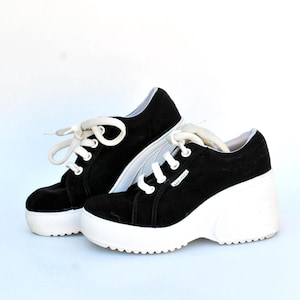 Op de afbeelding: Een paar zwarte platform sneakers met witte veters en zolen. De schoenen hebben een suède-achtige textuur en een chunky, retro design. De sneakers staan tegen een effen witte achtergrond.