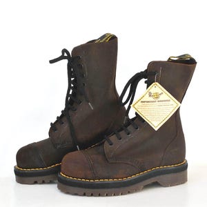 Pode incluir: Um par de botas de couro castanho escuro com atacadores pretos e solas grossas e tratoradas. As botas apresentam costuras amarelas ao longo da vira e uma presilha amarela e preta no calcanhar. Uma etiqueta bege com o texto Dr. Martens Important Warning está presa.