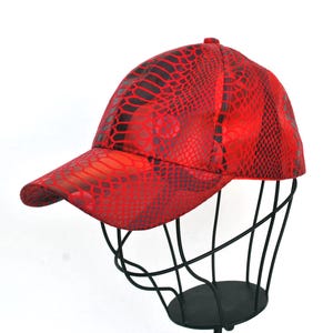 Può includere: Cappellino da baseball rosso con motivo a pelle di serpente. Il cappellino è esposto su una testa di manichino in filo di ferro nero su sfondo bianco. Ha una visiera curva e un bottone sulla parte superiore.