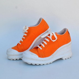 May include: A pair of bright orange canvas sneakers with white soles and laces. The shoes have a wedge heel and a classic sneaker design, perfect for casual wear.