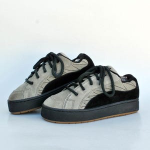May include: A pair of gray and black sneakers with black laces. The shoes have a suede-like texture and a thick black sole. The design includes geometric stitching details.