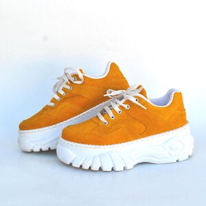 May include: A pair of mustard yellow platform sneakers with white soles and white laces. The chunky soles add height to the shoes. The sneakers are made of suede-like material.