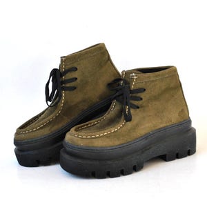 May include: A pair of olive green ankle boots featuring a suede-like texture, black laces, and thick black lug soles. The boots have a moccasin-style stitched toe box with contrasting light-coloured stitching along the seams and a high-top design.