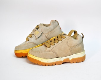 May include: A pair of beige suede sneakers with yellow accents and white soles. The sneakers have a lace-up closure and a zipper on the side.