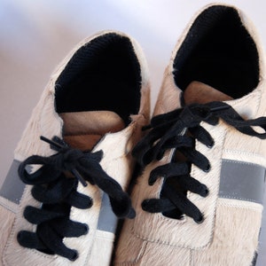 May include: A pair of white leather trainers with black laces and a grey stripe on each side. The shoes have a furry texture.