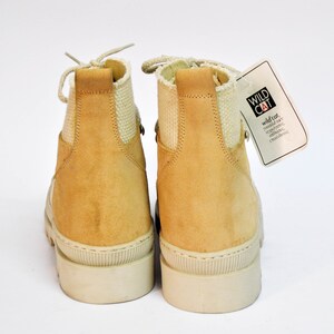 May include: A pair of tan suede ankle boots with white laces and a white rubber sole. The boots have a canvas panel on the back of the ankle. The boots are tagged with a white tag that says "WILD CAT" and "wild col. world star. independent. original. exclusive."