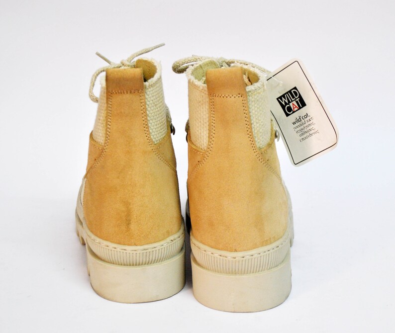 May include: A pair of tan suede ankle boots with white laces and a white rubber sole. The boots have a canvas panel on the back of the ankle. The boots are tagged with a white tag that says "WILD CAT" and "wild col. world star. independent. original. exclusive."