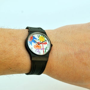 Puede incluir: Un reloj Swatch negro con una esfera blanca y un dise&ntilde;o colorido. La esfera del reloj presenta una flor, un p&aacute;jaro y una nube.