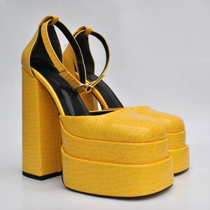 May include: A pair of yellow platform heels with ankle straps. The heels are made of a textured material that resembles crocodile skin.