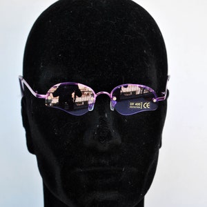 May include: Rimless oval sunglasses with a light purple frame and lenses. The lenses have a slight tint and reflect the surrounding environment. The sunglasses are displayed on a black mannequin head. The text "UV 400 PROTECTION" and "CE" are visible on the lens.