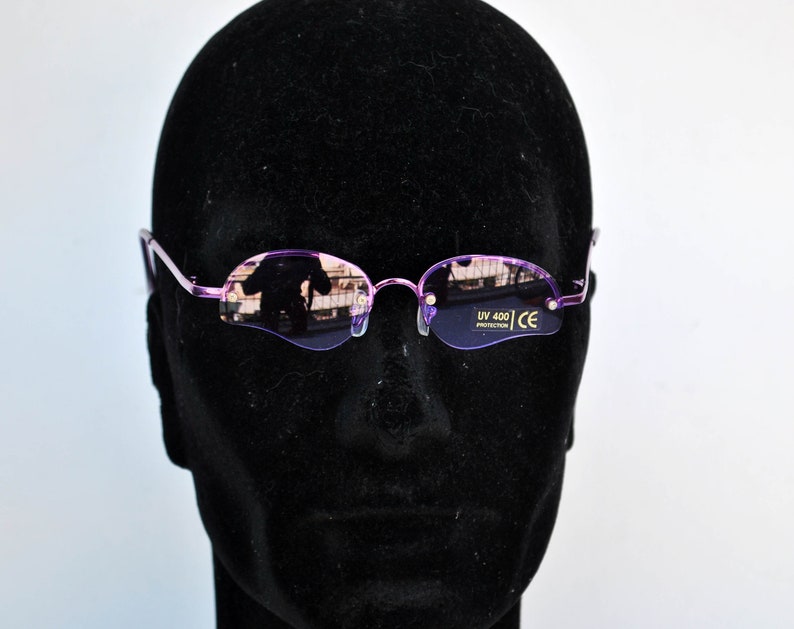 May include: Rimless oval sunglasses with a light purple frame and lenses. The lenses have a slight tint and reflect the surrounding environment. The sunglasses are displayed on a black mannequin head. The text "UV 400 PROTECTION" and "CE" are visible on the lens.