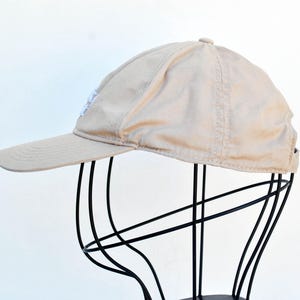 May include: A beige baseball cap with a white logo on the front. The cap is on a black stand.