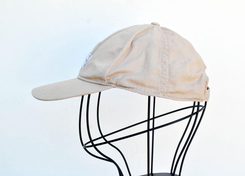 May include: A beige baseball cap with a white logo on the front. The cap is on a black stand.