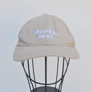 May include: A beige baseball cap with white embroidered text that reads "thanku, next".