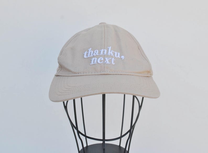 May include: A beige baseball cap with white embroidered text that reads "thanku, next".