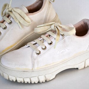 K&ouml;nnte beinhalten: Ein Paar wei&szlig;e Canvas-Sneaker mit dicken wei&szlig;en Sohlen und Schn&uuml;rsenkeln. Die Sneaker haben ein rosafarbenes Futter und ein dezentes Logo an der Seite.