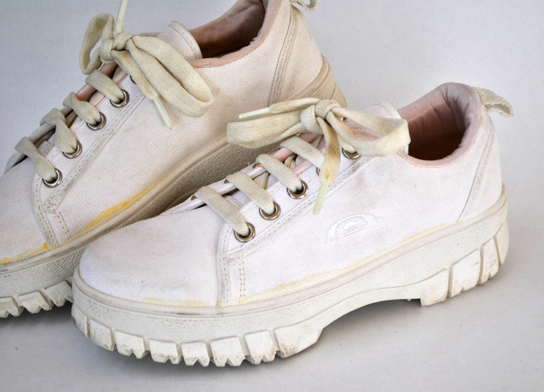 K&ouml;nnte beinhalten: Ein Paar wei&szlig;e Canvas-Sneaker mit dicken wei&szlig;en Sohlen und Schn&uuml;rsenkeln. Die Sneaker haben ein rosafarbenes Futter und ein dezentes Logo an der Seite.