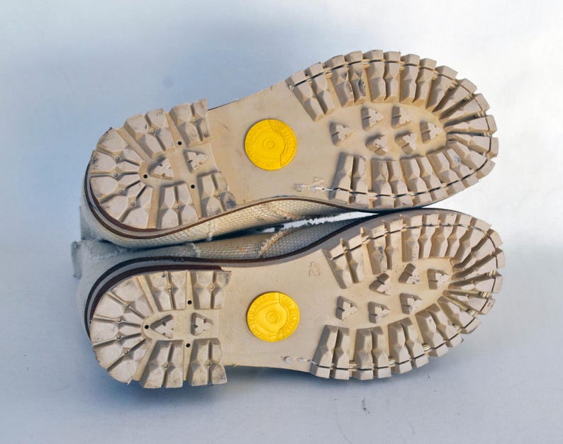 May include: A pair of white canvas shoes with brown rubber soles. The soles have a textured pattern and a yellow circle on each shoe.