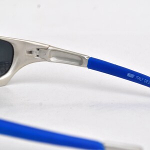 May include: Silver-toned sunglasses with blue rubber temple tips. The temple tips have the text "MADE ITALY DESIGN" printed on them.