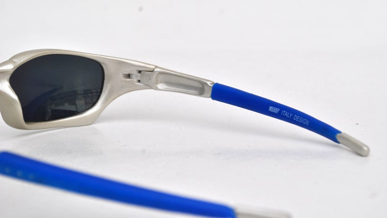 May include: Silver-toned sunglasses with blue rubber temple tips. The temple tips have the text "MADE ITALY DESIGN" printed on them.