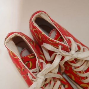 K&ouml;nnte beinhalten: Ein Paar rote High-Top-Sneaker mit wei&szlig;en Schn&uuml;rsenkeln und einem gold-roten Muster. Die Sneaker haben ein Obermaterial aus Canvas und eine Gummisohle.