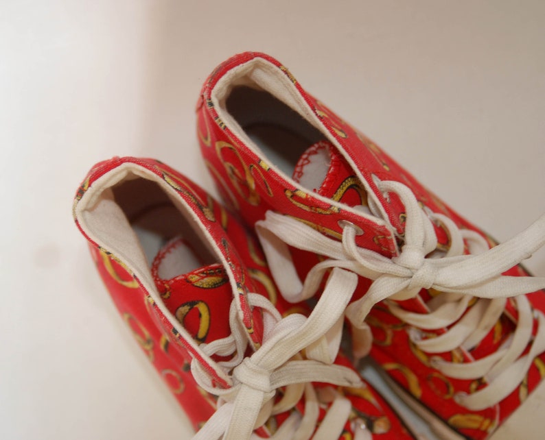 K&ouml;nnte beinhalten: Ein Paar rote High-Top-Sneaker mit wei&szlig;en Schn&uuml;rsenkeln und einem gold-roten Muster. Die Sneaker haben ein Obermaterial aus Canvas und eine Gummisohle.