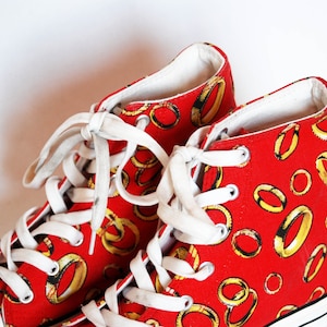 K&ouml;nnte beinhalten: Rote High-Top-Sneaker mit wei&szlig;en Schn&uuml;rsenkeln und einer wei&szlig;en Gummisohle. Die Sneaker sind mit einem Muster aus goldenen Ringen bedeckt.