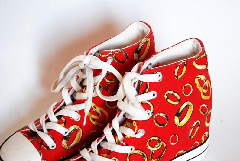 K&ouml;nnte beinhalten: Rote High-Top-Sneaker mit wei&szlig;en Schn&uuml;rsenkeln und einer wei&szlig;en Gummisohle. Die Sneaker sind mit einem Muster aus goldenen Ringen bedeckt.