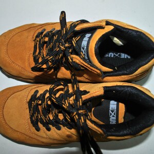 May include: A pair of brown suede hiking boots with black laces and a black and yellow logo that says "NEXUS".