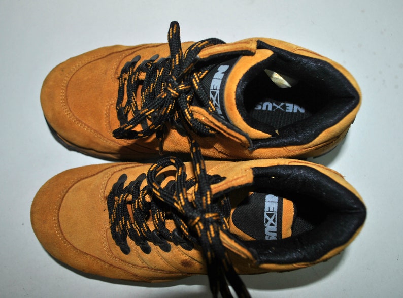 May include: A pair of brown suede hiking boots with black laces and a black and yellow logo that says "NEXUS".