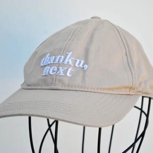 May include: A beige baseball cap with white embroidered text that reads "thanku, next".