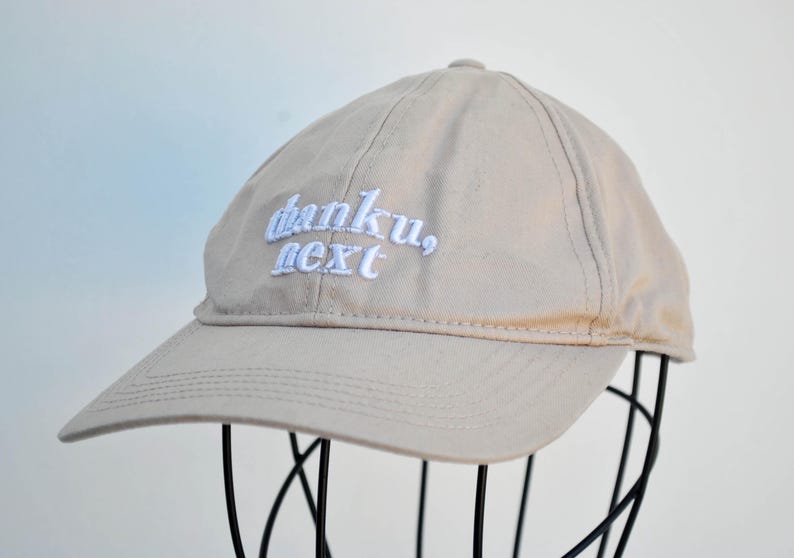 May include: A beige baseball cap with white embroidered text that reads "thanku, next".