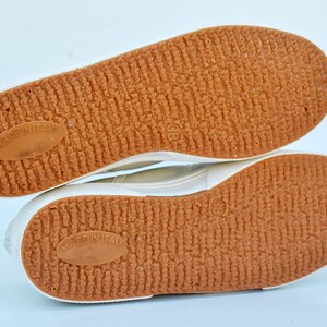 May include: Close-up of the bottom of a pair of white canvas shoes with brown rubber soles. The soles have a textured pattern and the words "Made in Italy" are printed on each sole.