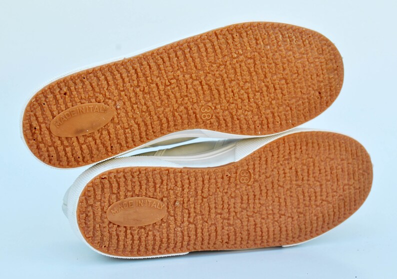 May include: Close-up of the bottom of a pair of white canvas shoes with brown rubber soles. The soles have a textured pattern and the words "Made in Italy" are printed on each sole.