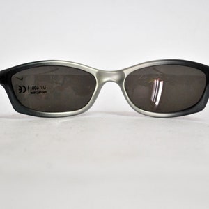 May include: Gray and black sunglasses with a silver frame and black arms. The sunglasses have a CE mark and the text "004 VU" on the right lens.