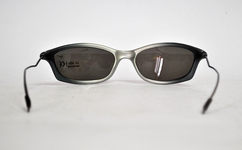 May include: Gray and black sunglasses with a silver frame and black arms. The sunglasses have a CE mark and the text "004 VU" on the right lens.