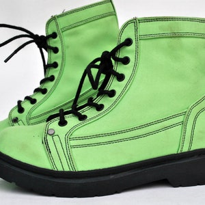 Op de afbeelding: Een paar felgroene combat boots met zwarte veters en zolen. De laarzen hebben een canvas-achtige textuur en zwarte stikseldetails. De veters zijn op een klassieke manier gestrikt. Een opvallend mode-item.