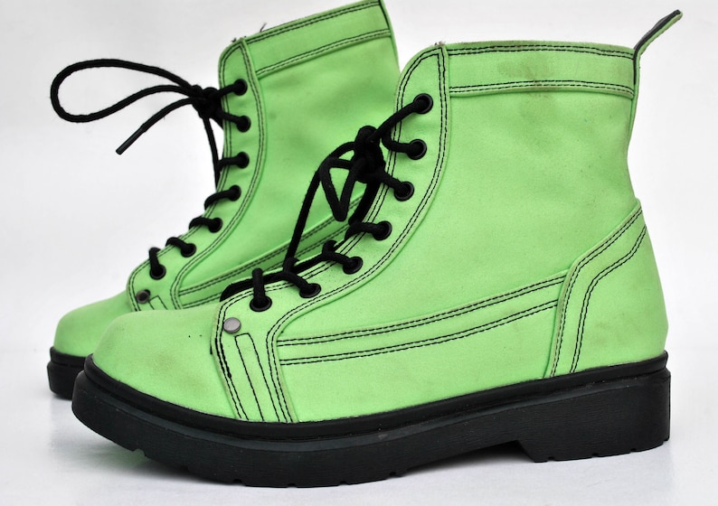 Op de afbeelding: Een paar felgroene combat boots met zwarte veters en zolen. De laarzen hebben een canvas-achtige textuur en zwarte stikseldetails. De veters zijn op een klassieke manier gestrikt. Een opvallend mode-item.