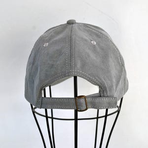 May include: A gray suede baseball cap with a brown adjustable strap. The cap is on a black display stand.
