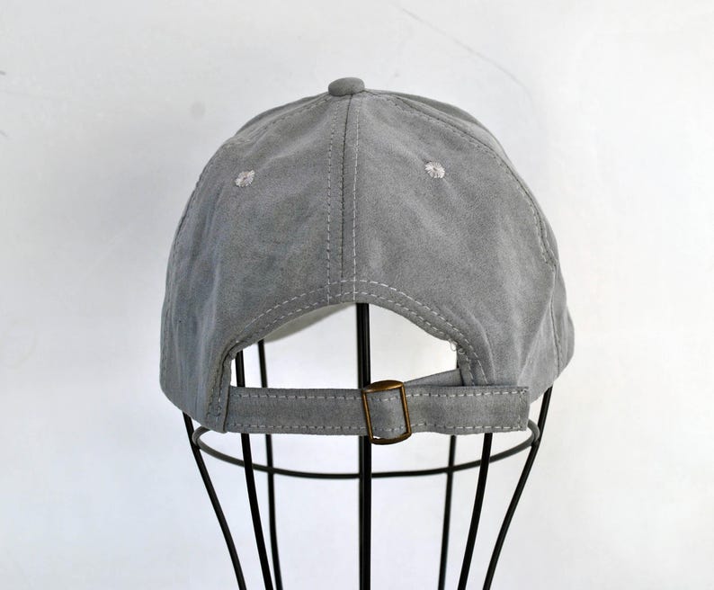 May include: A gray suede baseball cap with a brown adjustable strap. The cap is on a black display stand.