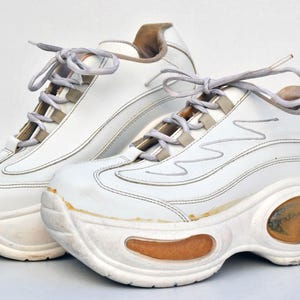 Puede incluir: Un par de zapatillas blancas de plataforma con suela gruesa y cierre de cordones. Las zapatillas tienen un aspecto ligeramente desgastado, con algunas rozaduras y marcas en el cuero.
