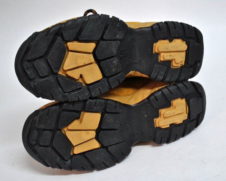 May include: Close-up of the soles of a pair of tan leather boots with black rubber outsoles. The soles have a textured pattern and the text "Made in Italy" is visible on the right boot.